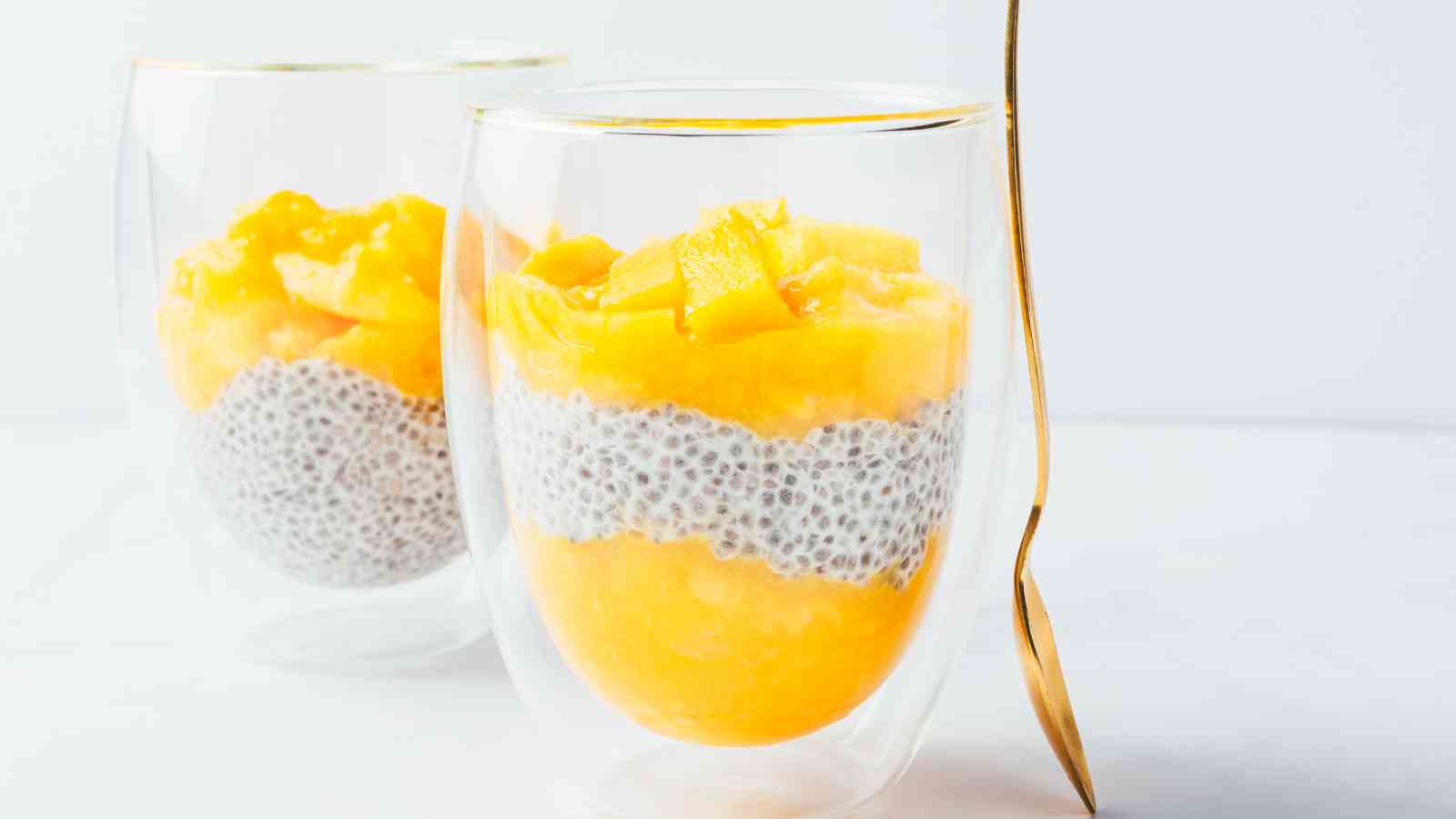 Two glass cups filled with mango chunks, chia seed pudding, and mango puree, with a golden spoon leaning against one cup, on a white surface.