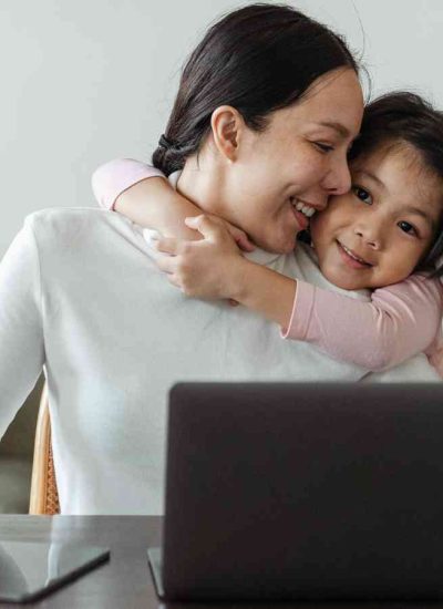 A woman using a tablet next to a laptop while a child hugs her from behind, both smiling. A mug is on the table, and a sofa and plants are in the background.