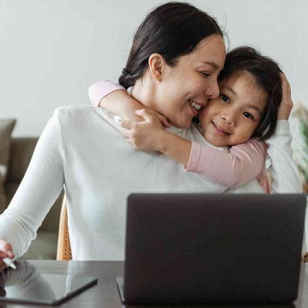A woman using a tablet next to a laptop while a child hugs her from behind, both smiling. A mug is on the table, and a sofa and plants are in the background.