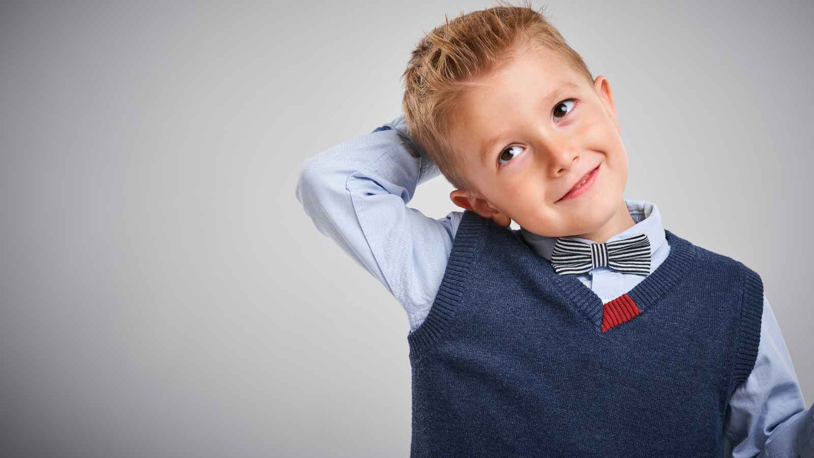 Young boy wearing a bow tie and sweater vest, looking up with one hand on his head, against a gray background.