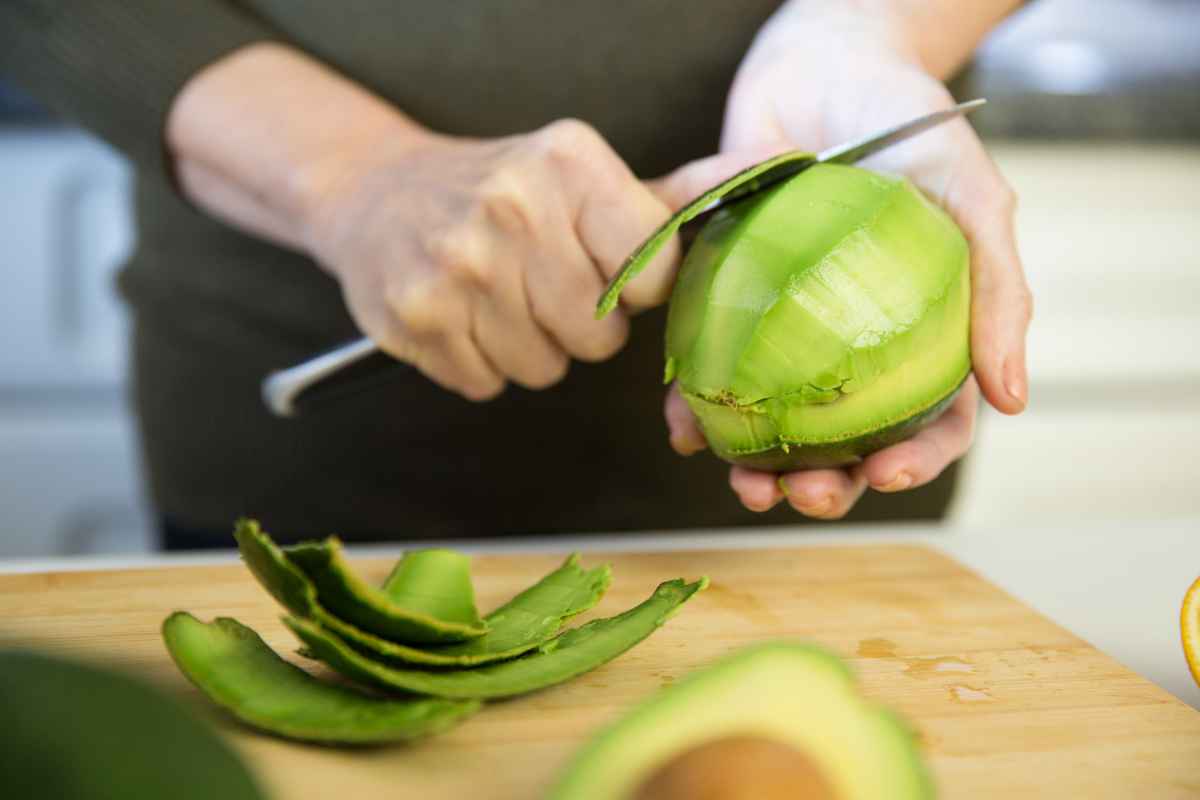 Person slicing an avocado with a knife on a wooden cutting board, several avocado slices nearby.
