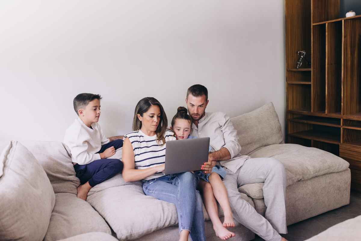 A family of four sits on a couch. The parents and two kids are looking at a laptop. They are in a living room with a wooden shelf.