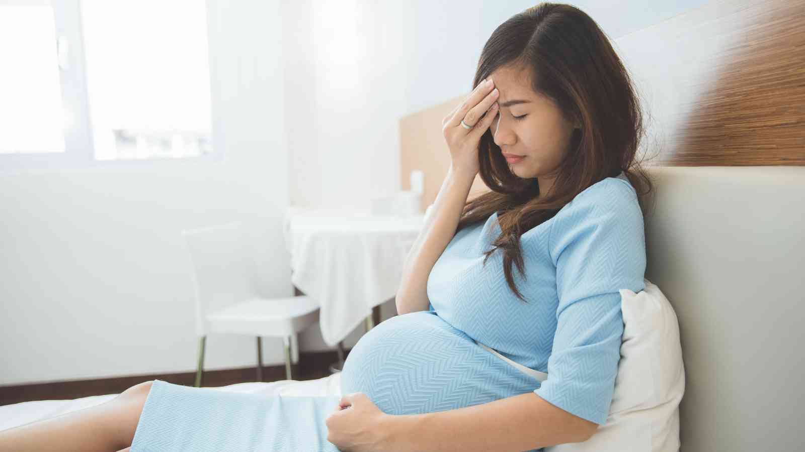 Pregnant woman in a blue dress sits on a bed, touching her forehead and appearing pensive or unwell, in a bright room with a table and chairs in the background.