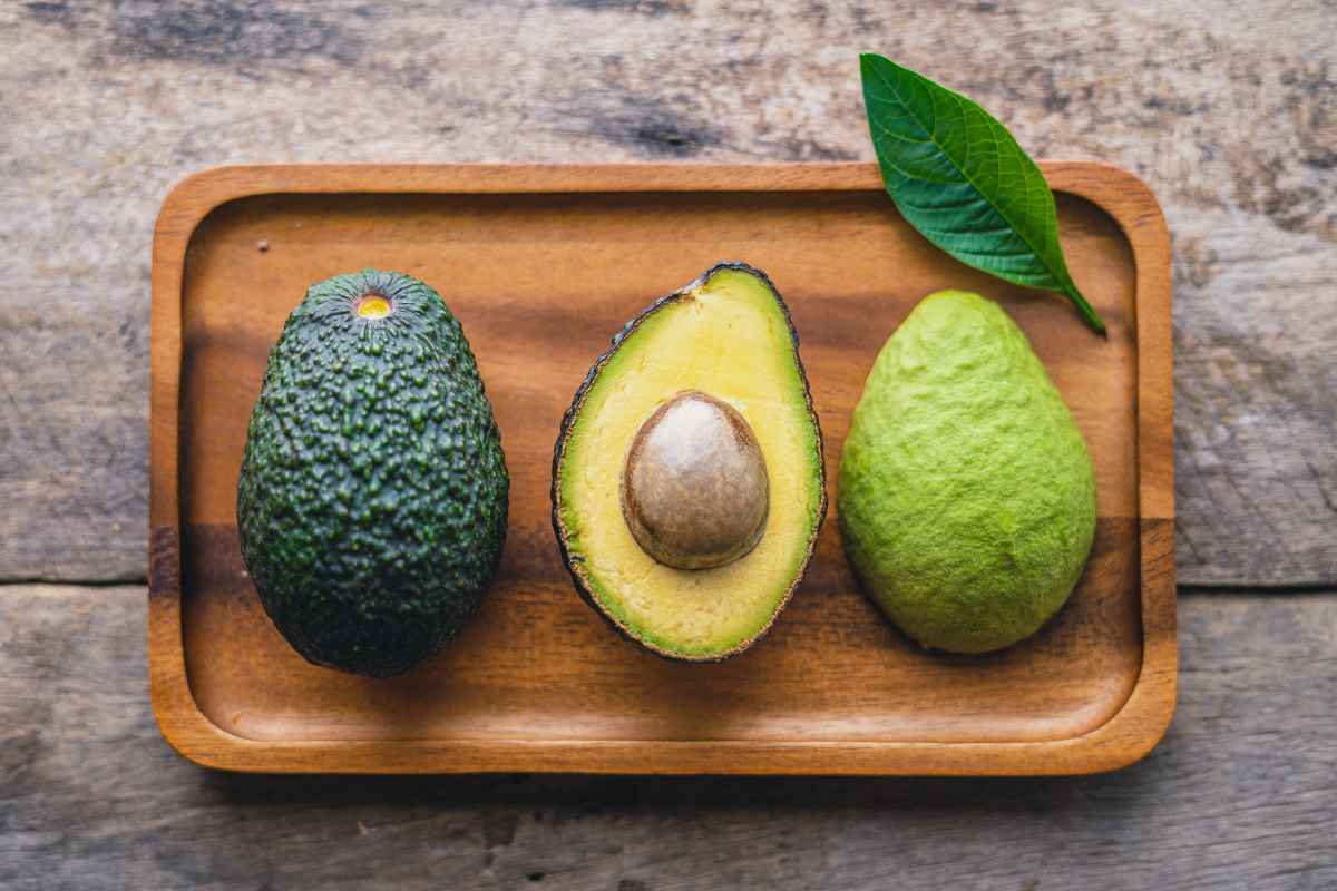 A wooden tray holds a whole avocado, a halved avocado with the pit, and a peeled avocado, with a leaf alongside, on a wooden surface.