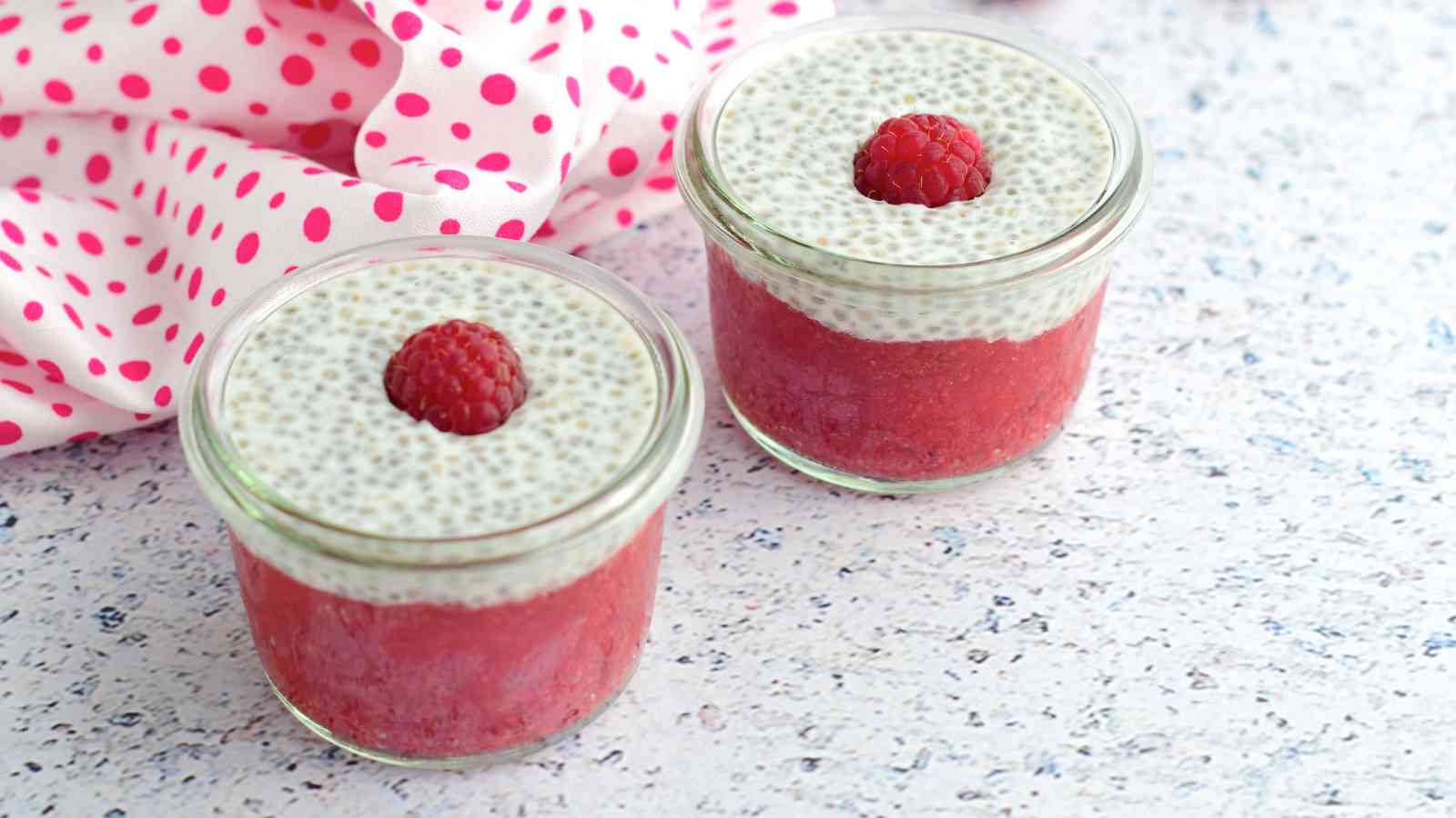Two jars of chia pudding with raspberry puree and topped with a raspberry are placed on a speckled surface with a pink polka dot cloth nearby.