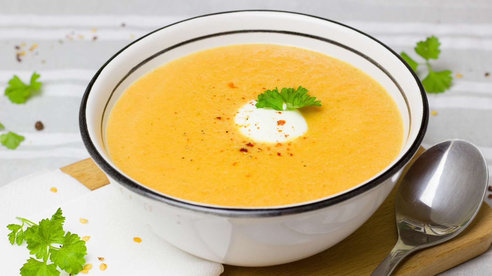 A bowl of creamy orange soup with a dollop of cream and a parsley garnish. A spoon and sprigs of parsley rest on a white napkin nearby.