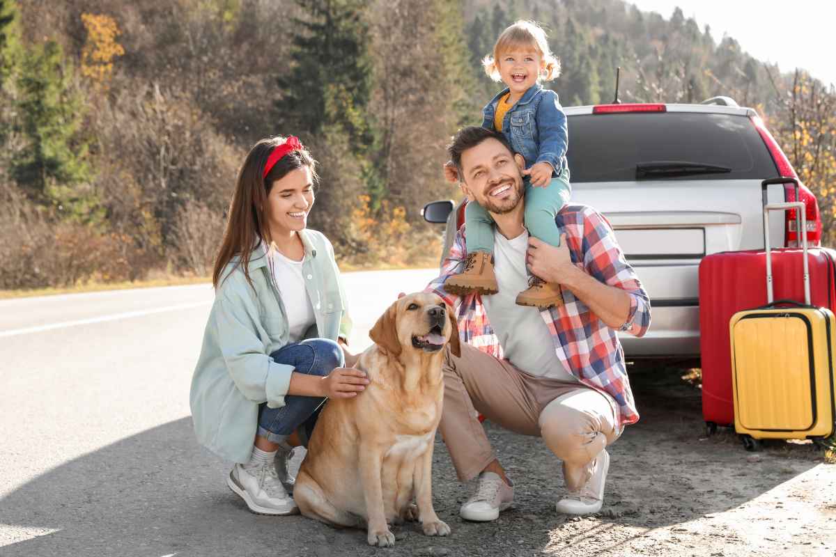 A family with a dog poses beside a parked car on a rural road. The man holds a child on his shoulders, and luggage is visible next to the car. Trees line the background.