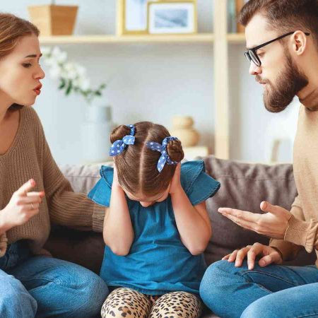 Two adults are sitting on a couch talking to a child who covers her ears, looking distressed. The setting is a living room with shelves in the background.