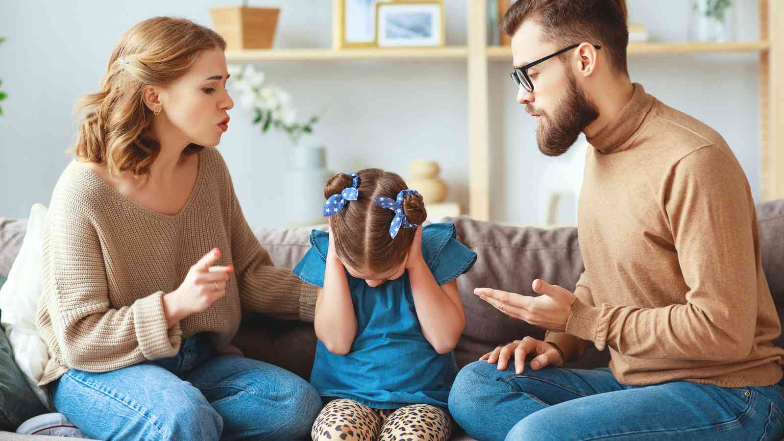 Two adults are sitting on a couch talking to a child who covers her ears, looking distressed. The setting is a living room with shelves in the background.