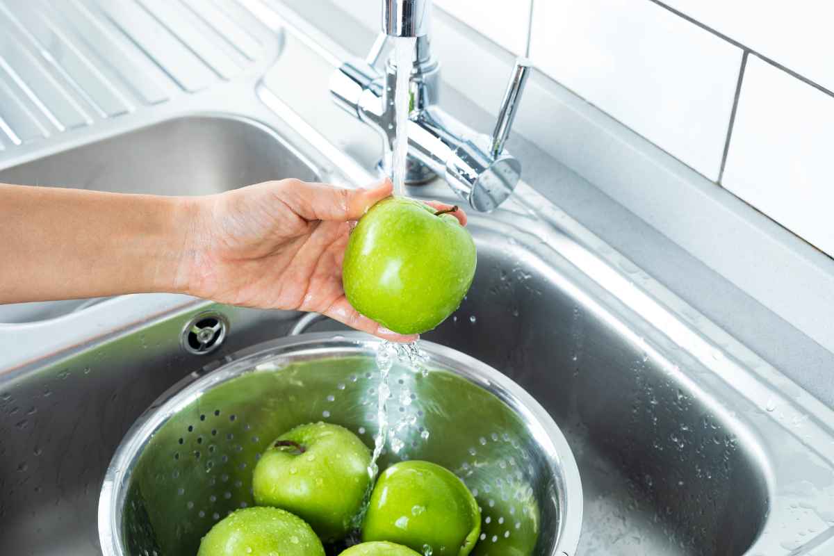 A hand washes a green apple under running water in a kitchen sink, with more green apples in a metal colander below.