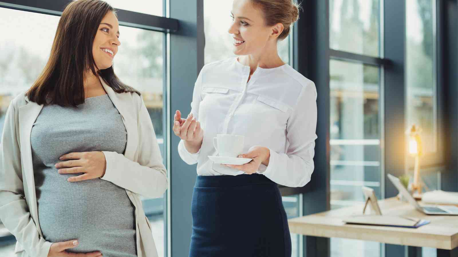 Two women standing indoors near windows. One is pregnant, wearing a gray dress and smiling. The other holds a cup and gestures, wearing a white blouse and dark skirt. They seem to be conversing.
