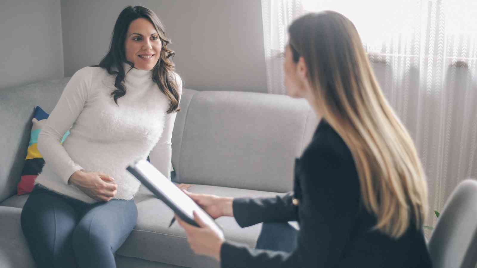Two women sit on a couch in conversation. One is holding a notebook, and the other appears to be pregnant, touching her belly.