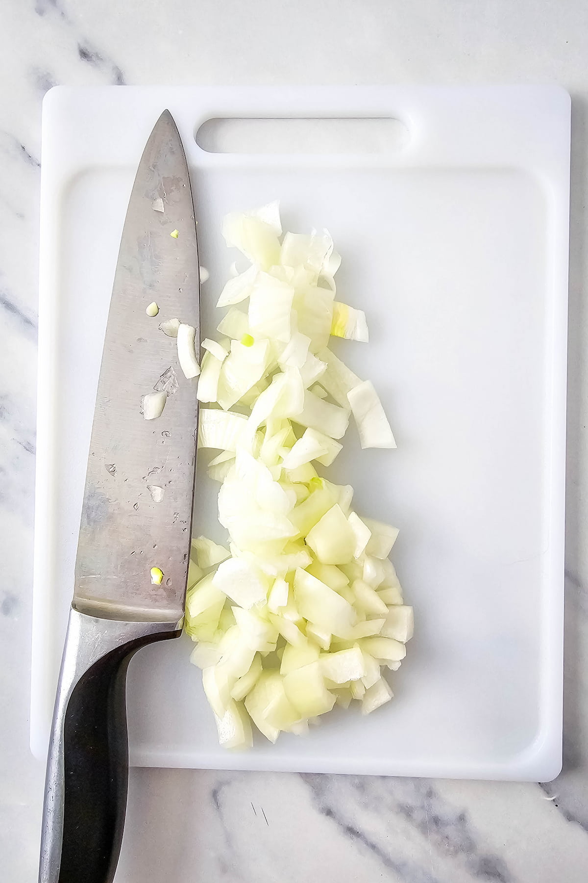 A chef's knife and chopped onions on a white cutting board are placed on a marble countertop.