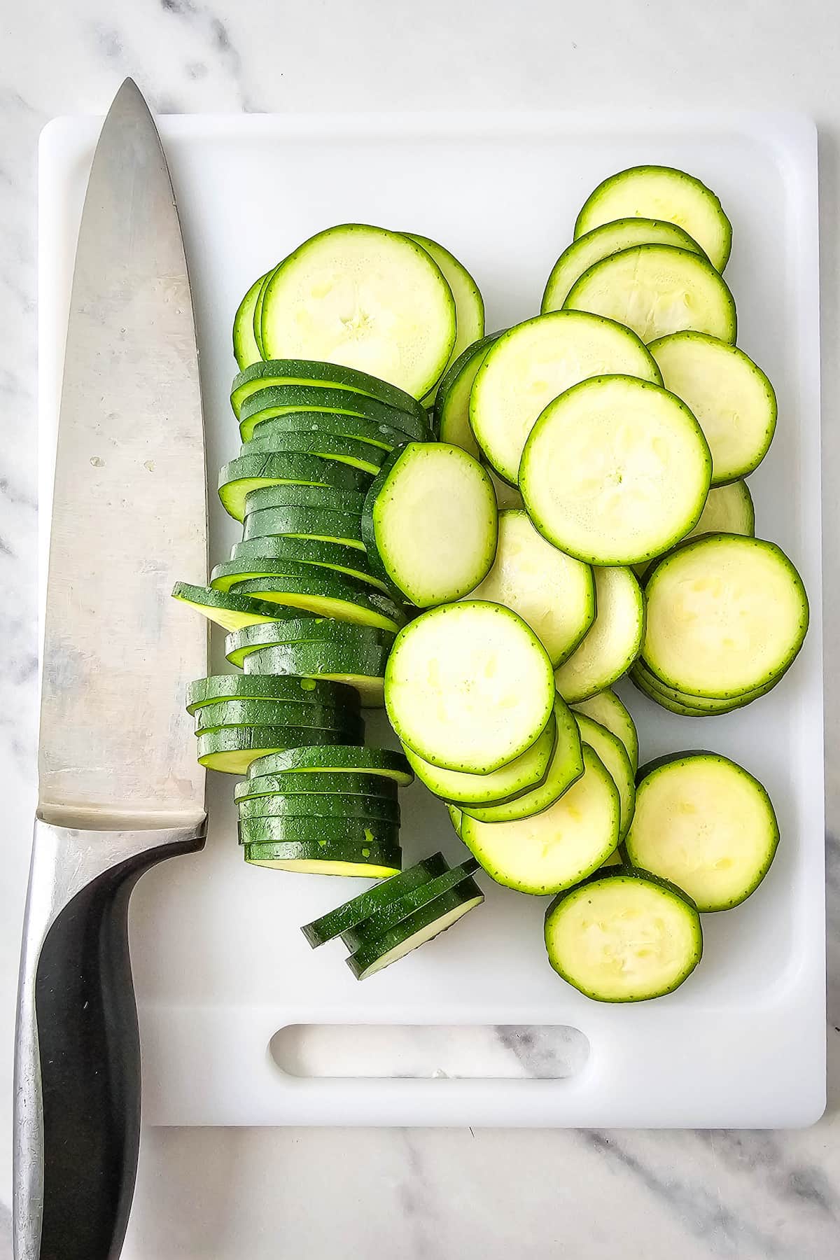 A chef’s knife and sliced zucchini are arranged on a white cutting board.