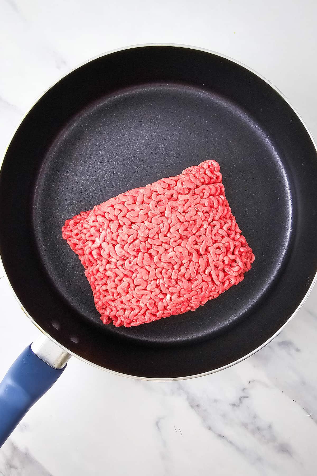 A block of raw ground beef sits in the center of a black nonstick frying pan on a marble countertop.