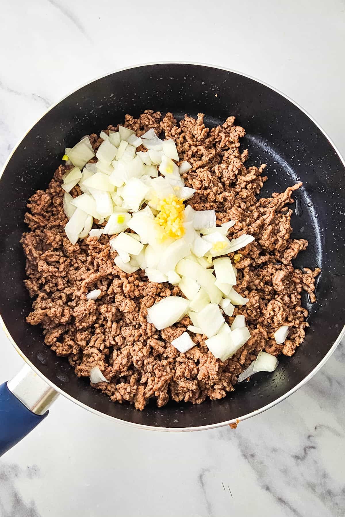 Ground beef cooks in a black skillet with chopped onions and minced garlic on top, set on a white marble countertop.