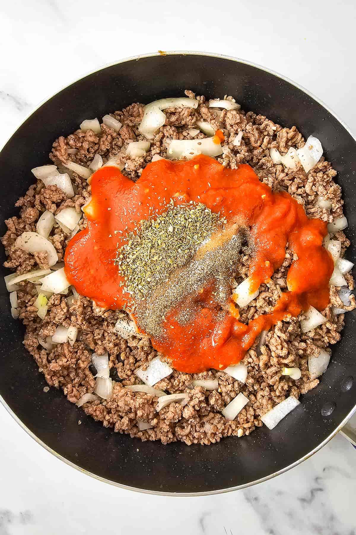 Ground beef and chopped onions cooking in a skillet, topped with marinara sauce and a sprinkle of dried herbs for a delicious ground beef and zucchini casserole.