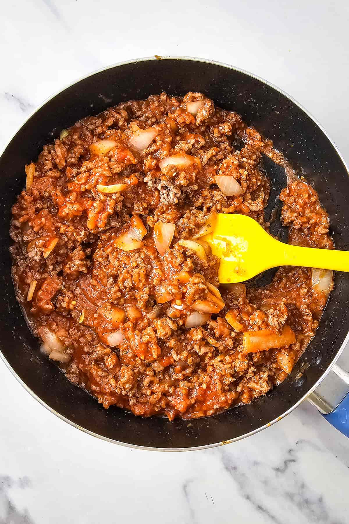 Ground beef, chopped onions, and sauce being stirred with a yellow spatula in a black skillet on a white surface.