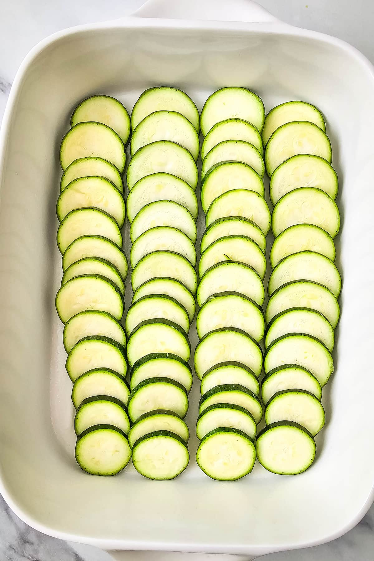 Sliced zucchini arranged in neat rows inside a white rectangular baking dish, perfect for assembling a hearty ground beef and zucchini casserole.