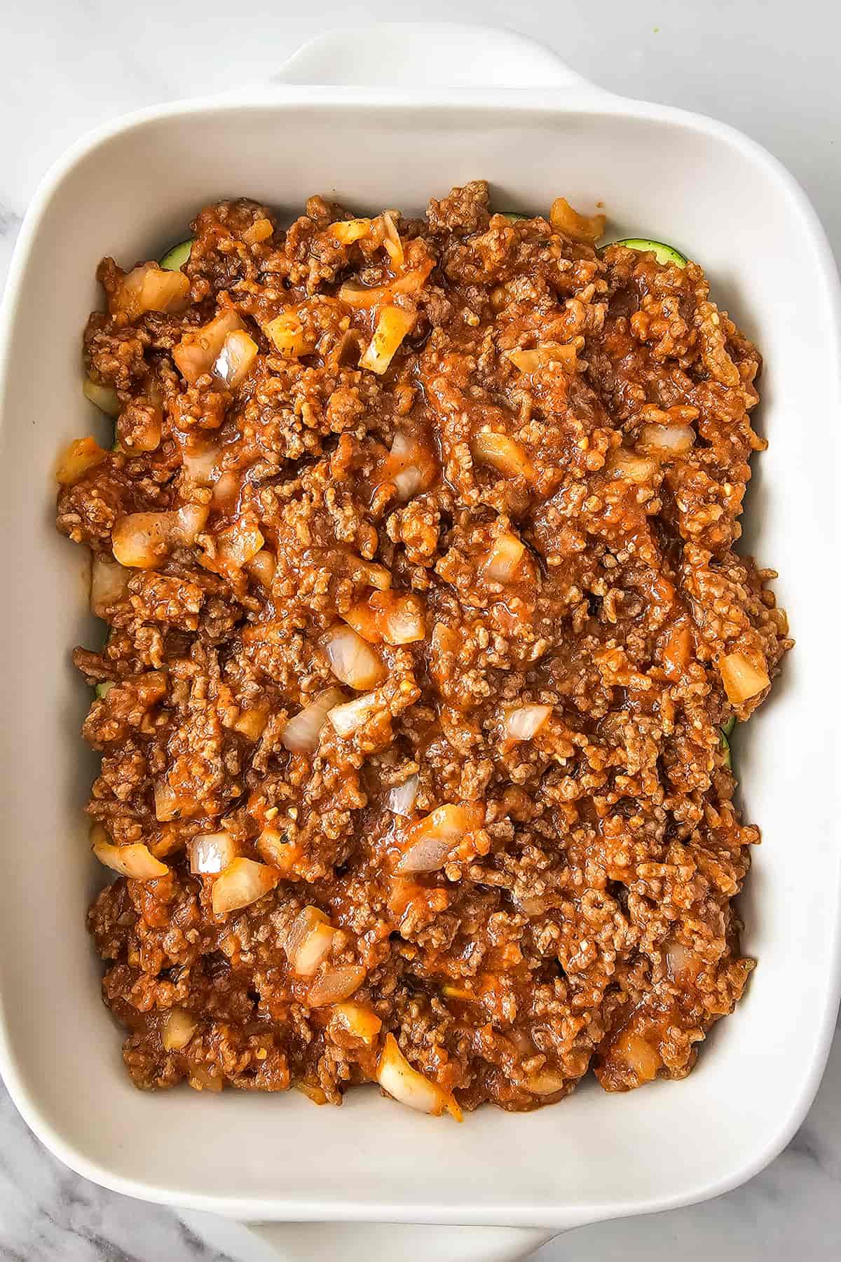 Ground beef and zucchini casserole featuring ground beef cooked with onions and marinara sauce, then spread evenly in a white rectangular baking dish.