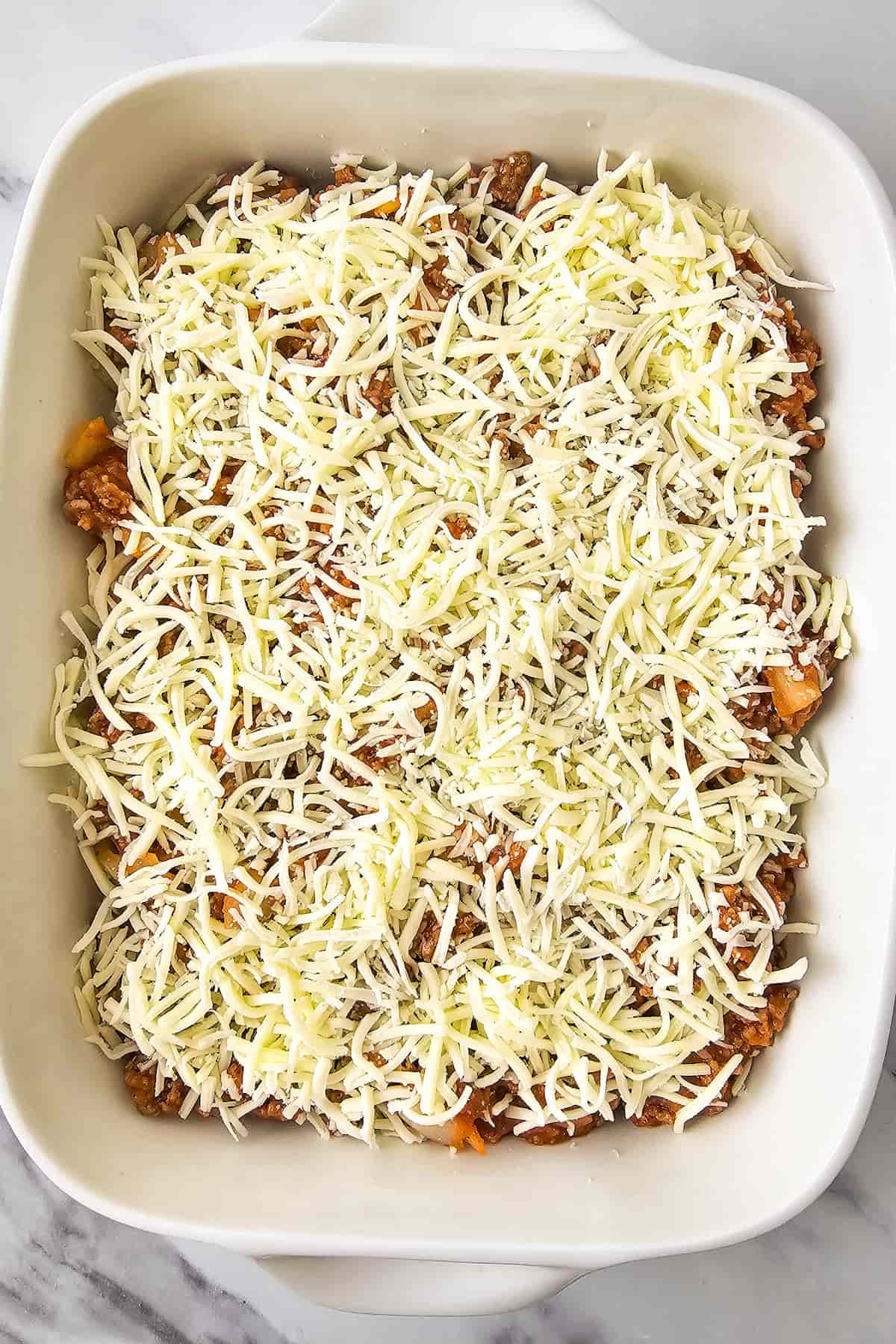A white baking dish filled with ground beef and zucchini casserole, topped with shredded cheese, ready to be baked.