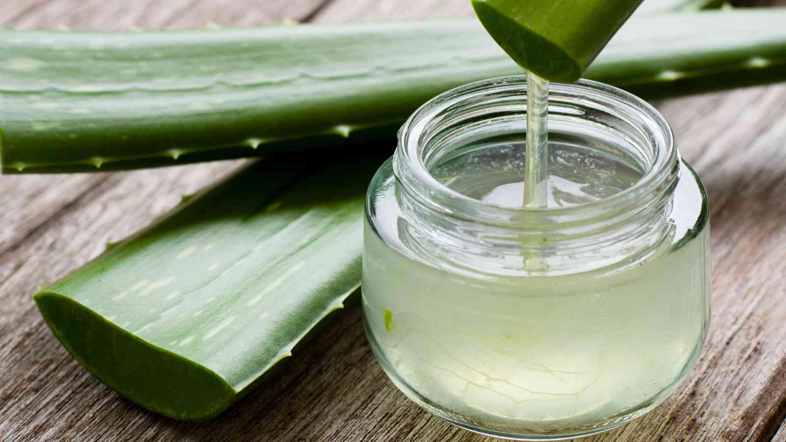 A jar of clear aloe vera gel with aloe leaves on a wooden surface, showing the gel being extracted from a cut leaf segment.