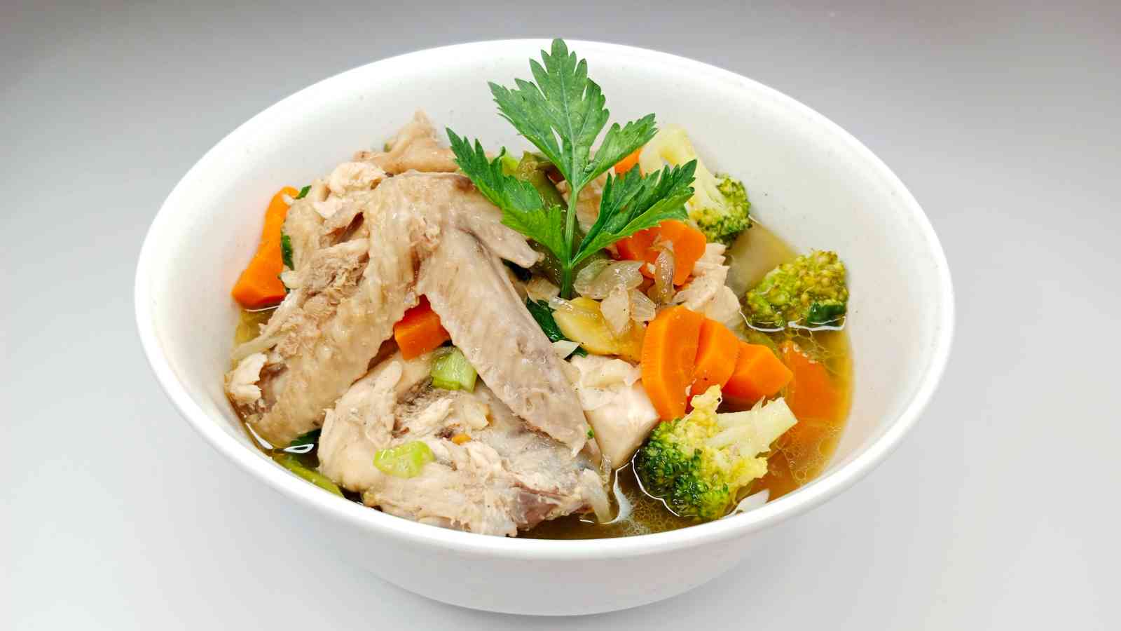 A bowl of chicken soup with sliced carrots, broccoli, and herbs, garnished with parsley.