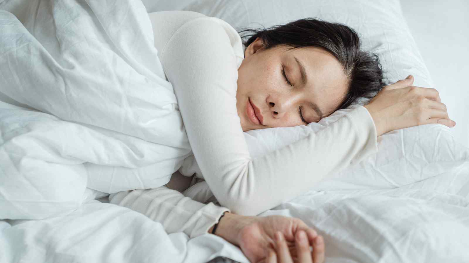 Person sleeping peacefully on a white bed, wrapped in a white blanket, with head resting on a white pillow.