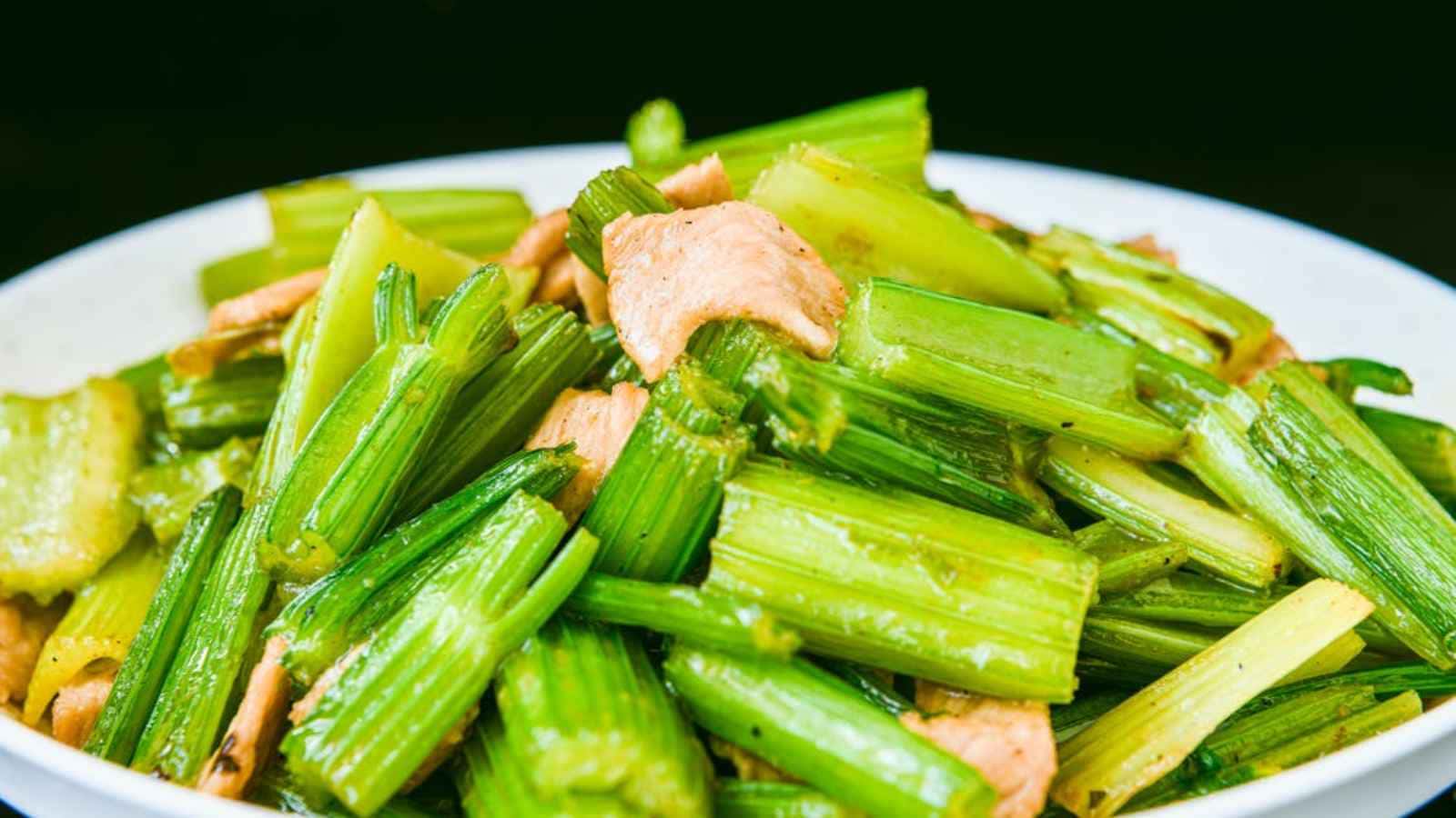 A dish of stir-fried celery and chicken pieces, served on a white plate against a dark background.