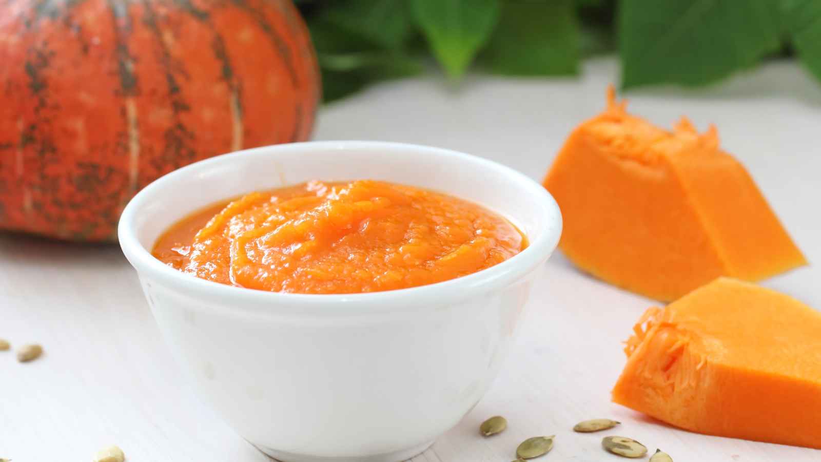 A white bowl of orange pumpkin puree sits on a white surface, with fresh pumpkin chunks and seeds nearby. A whole pumpkin is in the background.
