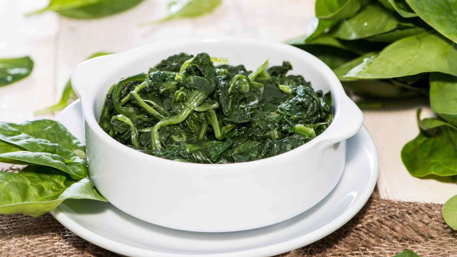 A white bowl filled with cooked spinach, placed on a white plate. Fresh spinach leaves scattered around on the table.