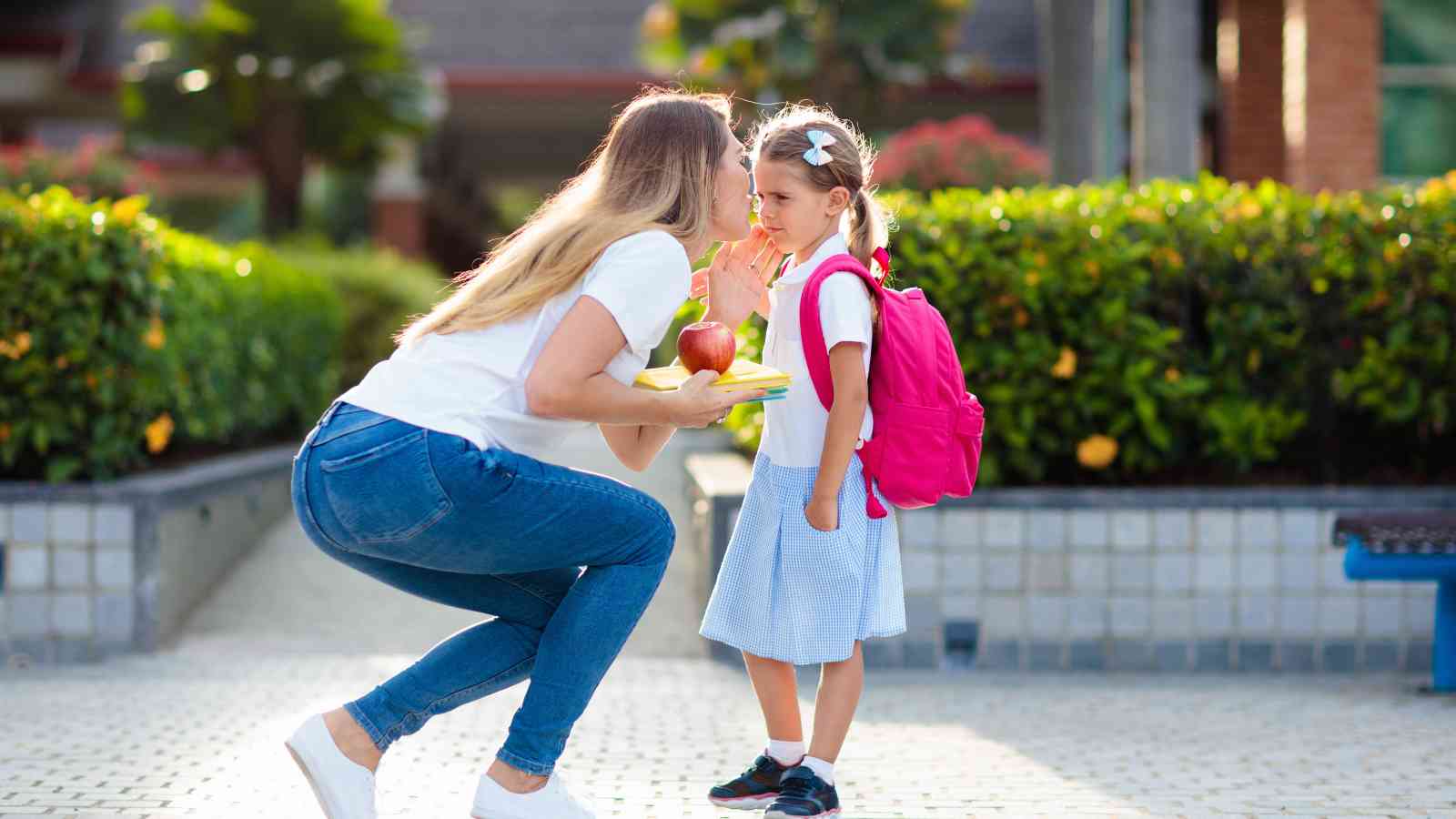A woman kneels in front of a young girl with a pink backpack, offering her an apple and plate, outdoors near greenery and buildings.