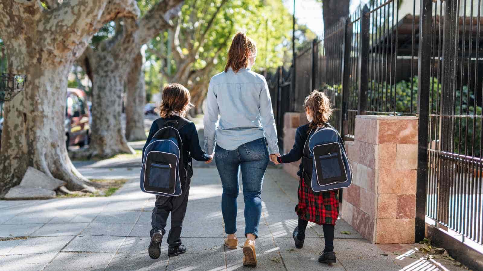 An adult walks on a sidewalk holding hands with two school-aged children wearing backpacks and uniforms, surrounded by trees and a fence.