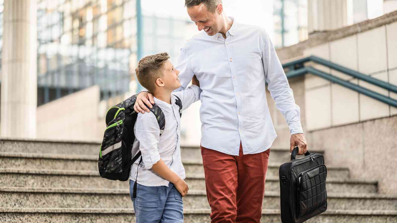 A man holding a briefcase walks down outdoor steps with a school-aged boy carrying a backpack. Both are smiling and looking at each other.