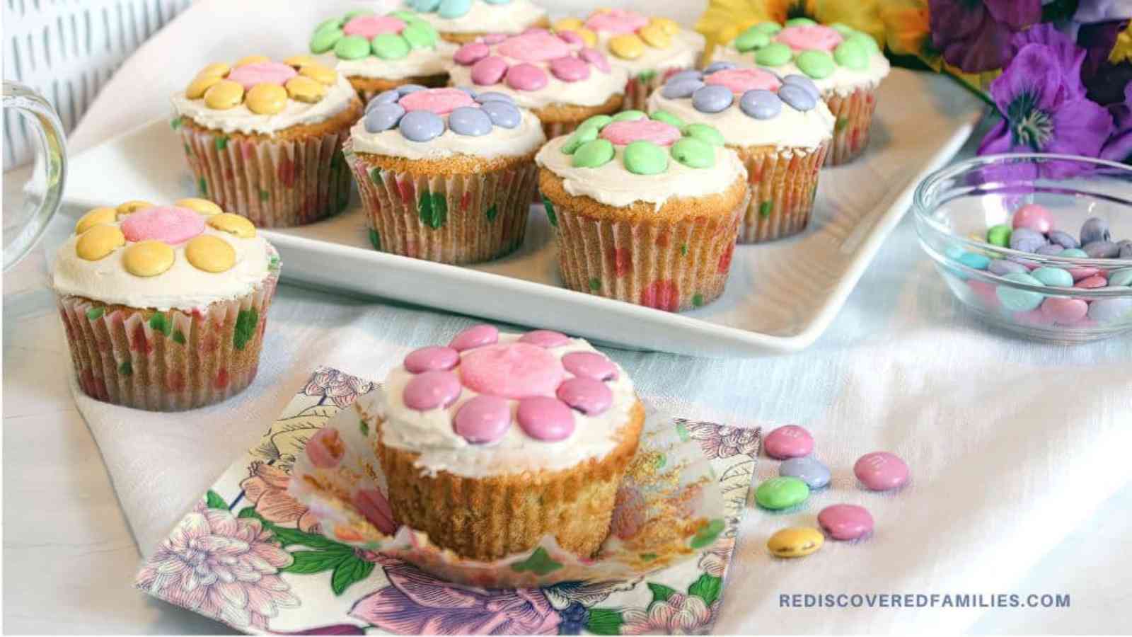 A platter of cupcakes decorated with pastel-colored candies to resemble flowers, with a single cupcake on a floral napkin in the foreground.
