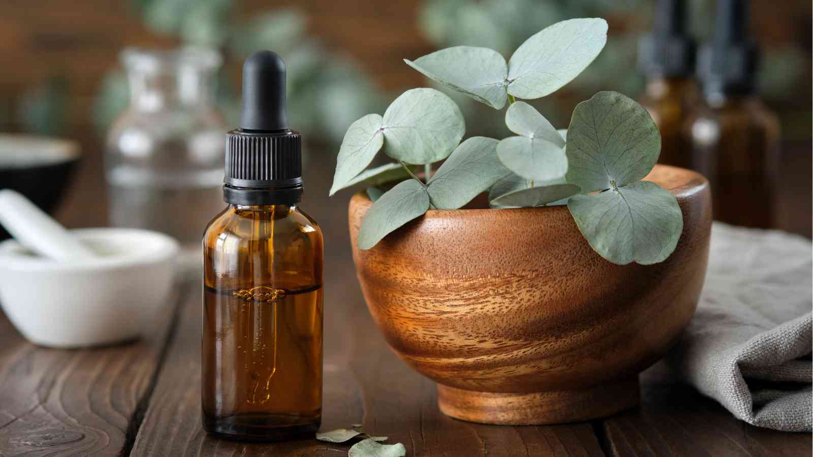 A brown dropper bottle sits next to a wooden bowl filled with eucalyptus leaves on a wooden table. Other glass bottles and a mortar and pestle are blurred in the background.