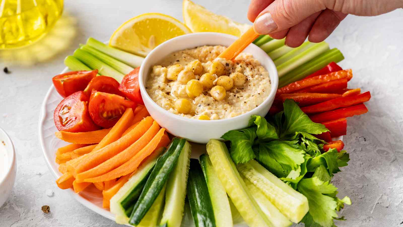 A hand dips a carrot stick into a bowl of hummus, surrounded by sliced cucumbers, celery, carrots, tomatoes, and bell peppers on a white plate.