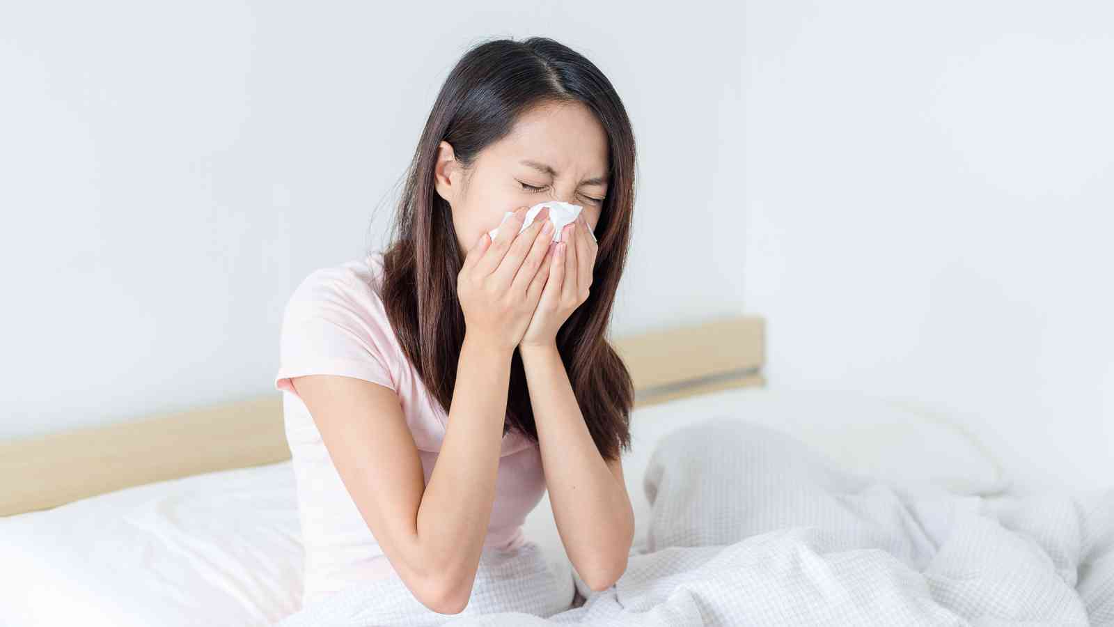 Person sitting on a bed, sneezing into a tissue.
