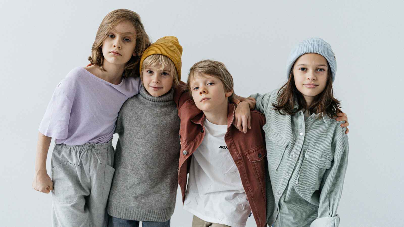 Four children stand closely together against a plain background, wearing casual clothing and beanies, with neutral expressions.