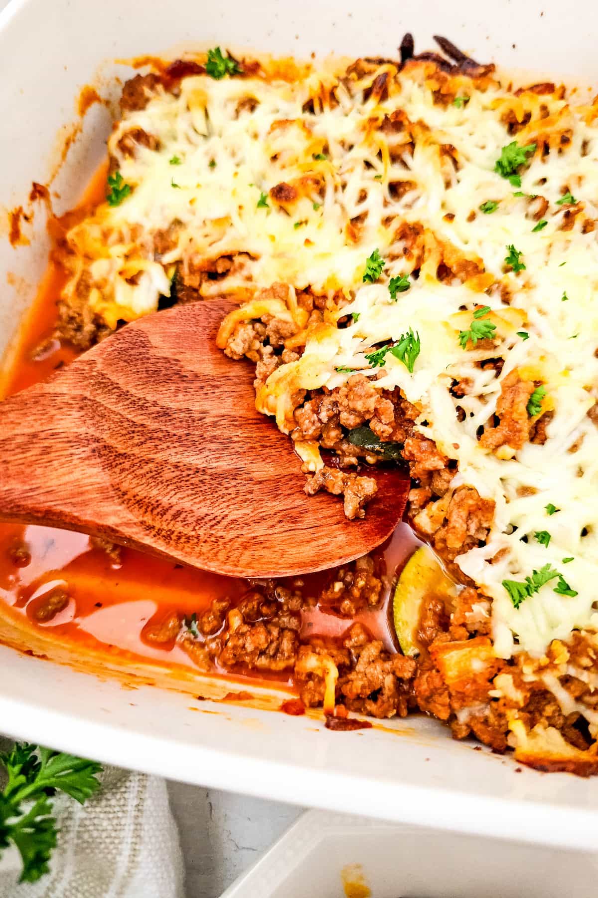 A wooden spatula scoops a portion of ground beef and zucchini casserole topped with melted cheese and herbs in a white dish.