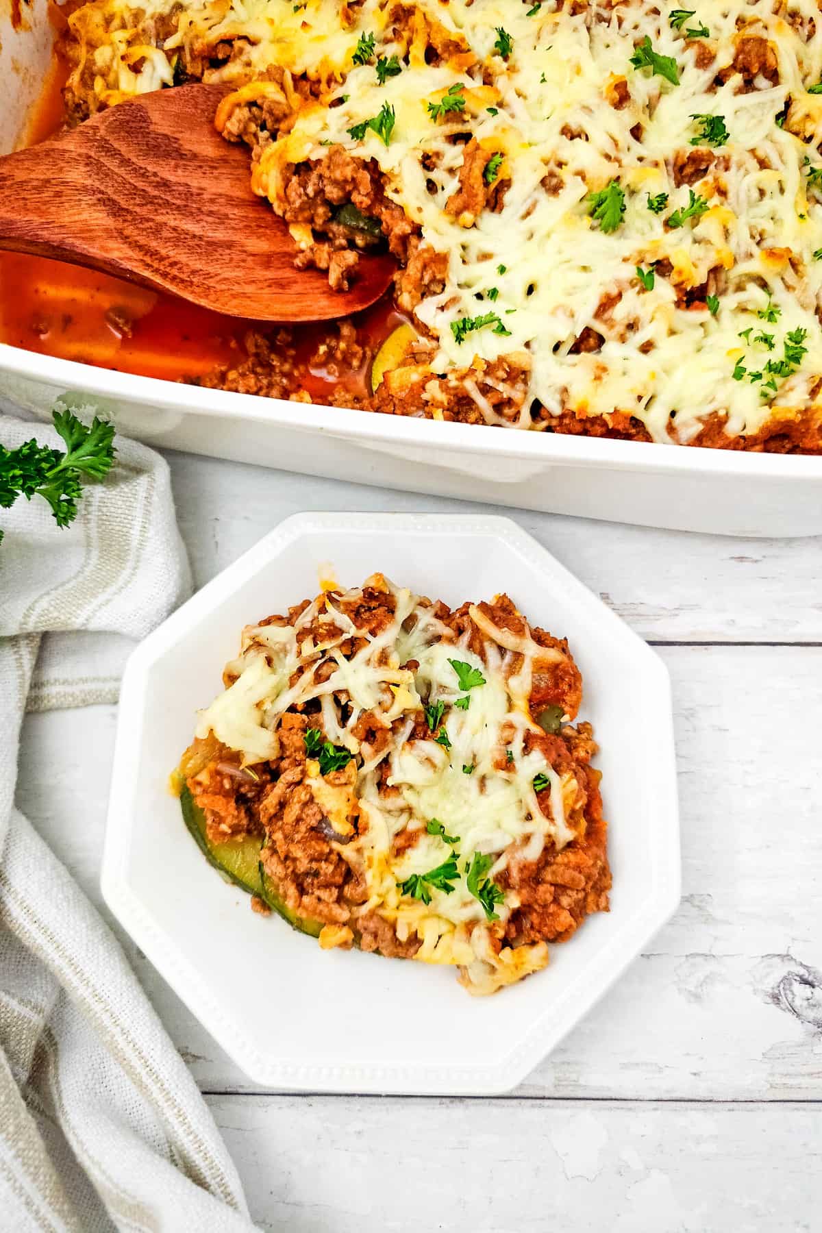 A serving of ground beef and zucchini casserole topped with melted cheese and parsley on a white plate, with the casserole dish full of savory ground beef and zucchini casserole in the background.