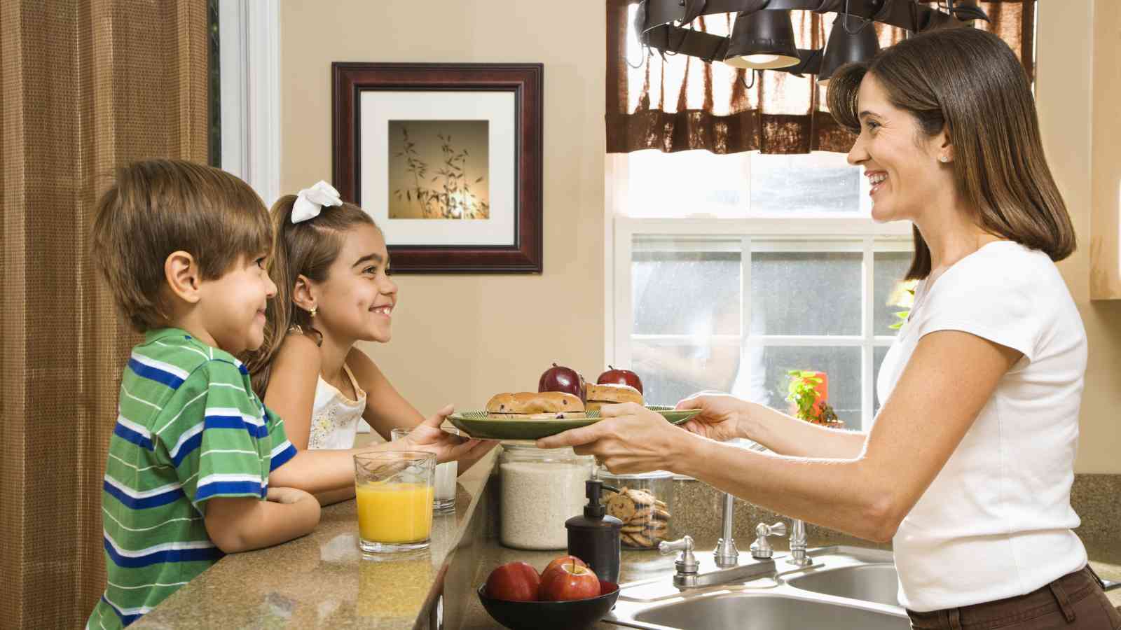 A woman hands a plate of cookies to two children in a kitchen, with a glass of orange juice on the counter.