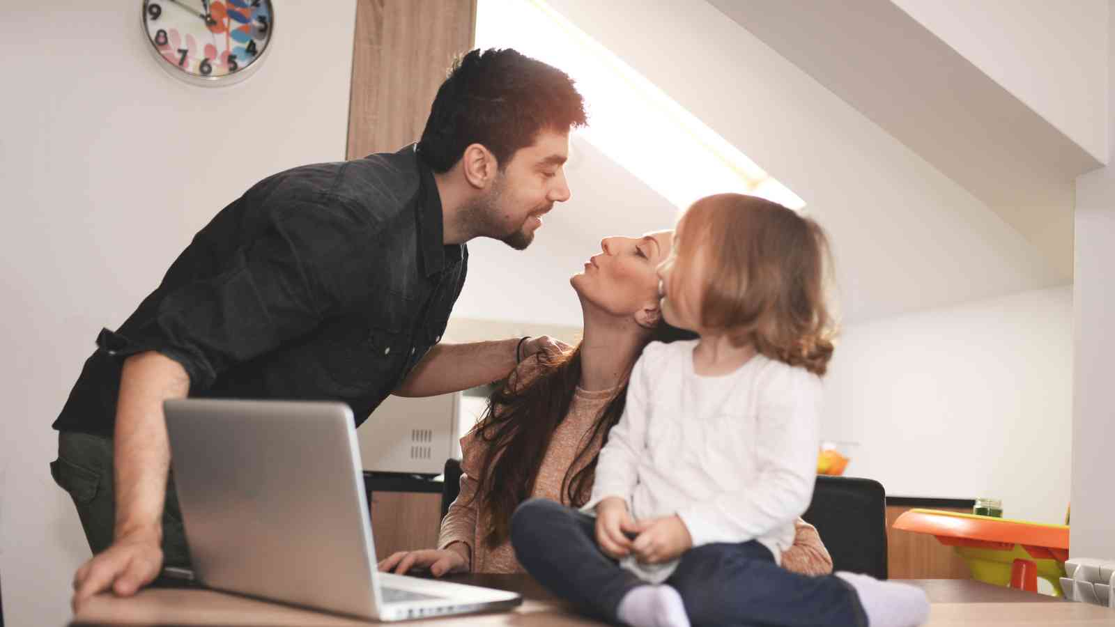 A couple exchanges a kiss while their child sits on a table beside a laptop in a cozy attic room.