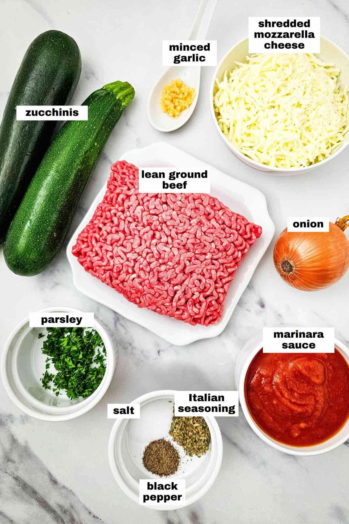 Ingredients for a delicious ground beef and zucchini casserole are displayed on a marble surface: zucchinis, minced garlic, shredded mozzarella cheese, lean ground beef, onion, parsley, salt, Italian seasoning, black pepper, and marinara sauce.