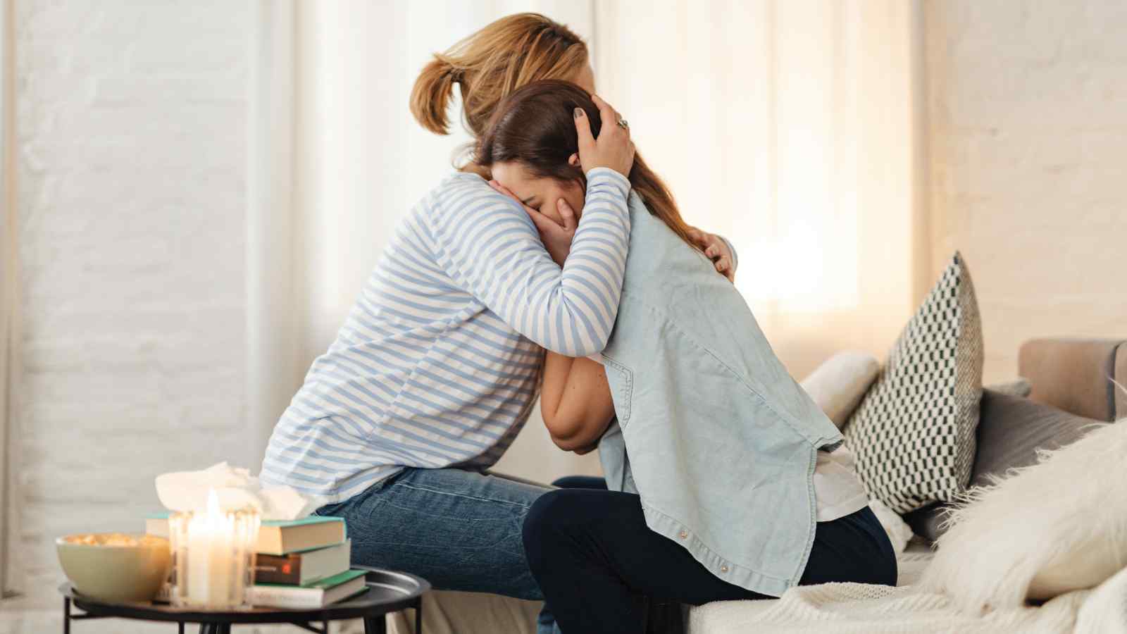 Two people sitting on a sofa share an emotional embrace. A side table with books and lit candles is next to them.