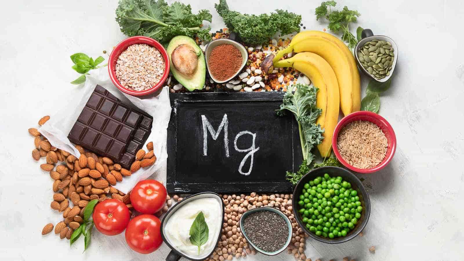 Various magnesium-rich foods, like almonds, bananas, chocolate, kale, peas, avocado, and grains, arranged around a chalkboard displaying the chemical symbol "Mg.