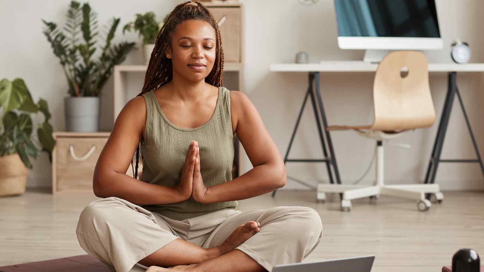 Person sitting cross-legged on the floor, eyes closed, and palms together in a meditation pose. Background includes a desk, chair, and plants.