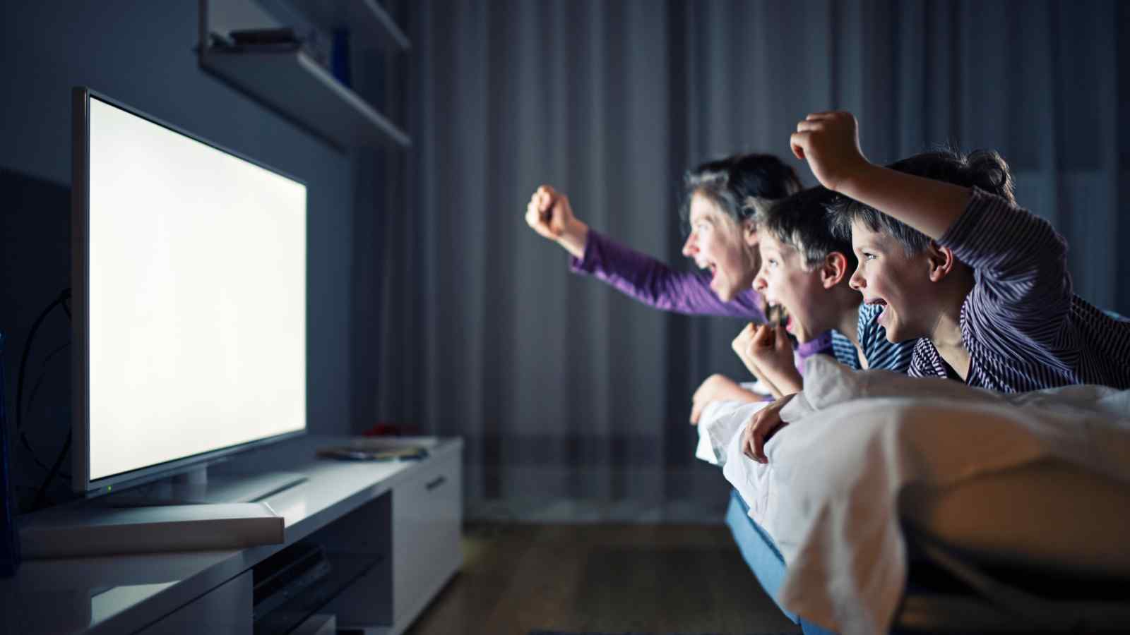 Three excited children watch a bright screen in a dimly lit room, cheering with raised arms while lying on a sofa.