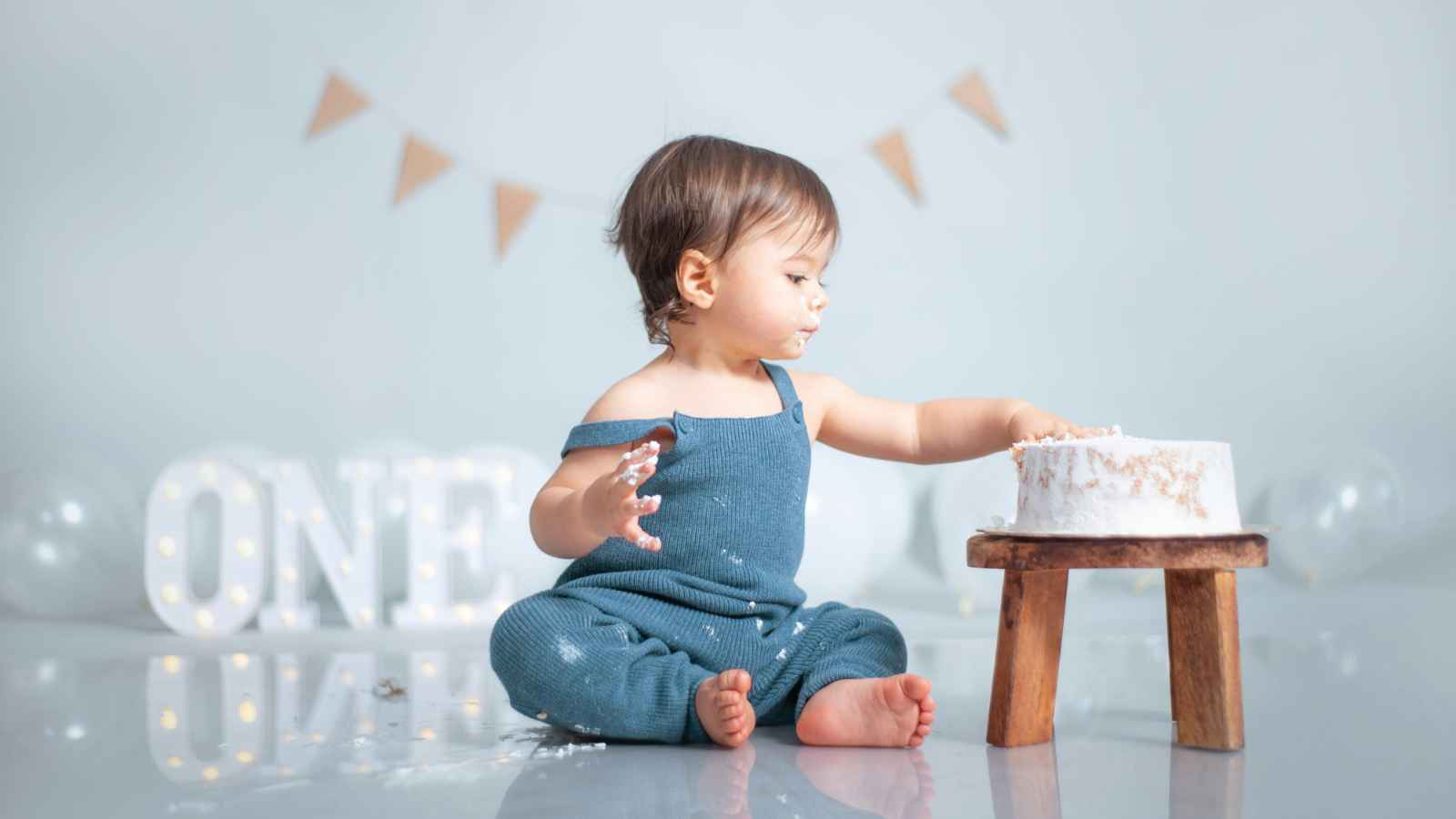 A toddler in a blue outfit sits on the floor touching a cake on a small stand. In the background, a banner and illuminated letters spell "ONE.