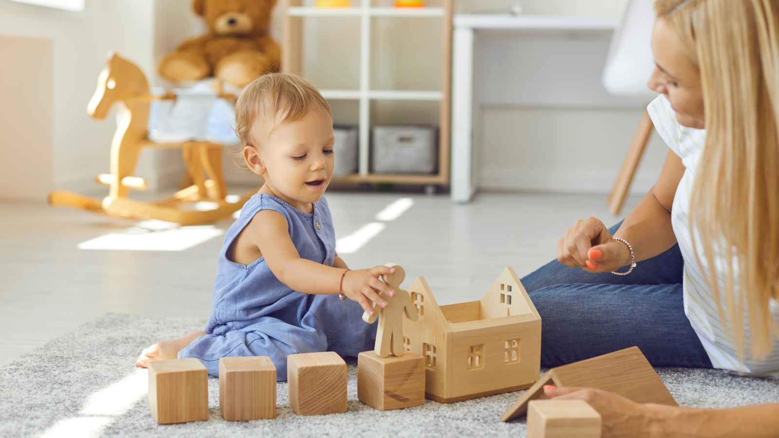 A toddler in a blue dress plays with wooden blocks and a toy house on a carpeted floor, while an adult nearby assists. A rocking horse and teddy bear are in the background.